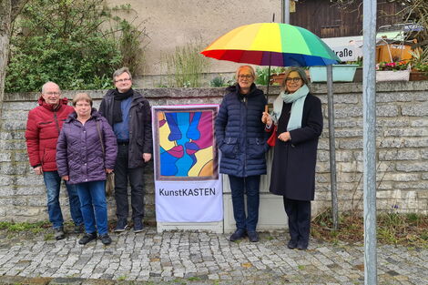 5 Personen zusammen vor einem zum Kunstkasten verhüllten Netzverteiler an einer Straße in Lichtenberg. 2 Personen stehen unter einem bunten großen Regenschirm.