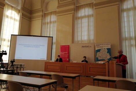 Pressekonferenz, 2 Männer und 1 Frau. Die Frau redet am Podest. Im Hintergrund wird eine Powerpointpräsentation gezeigt.